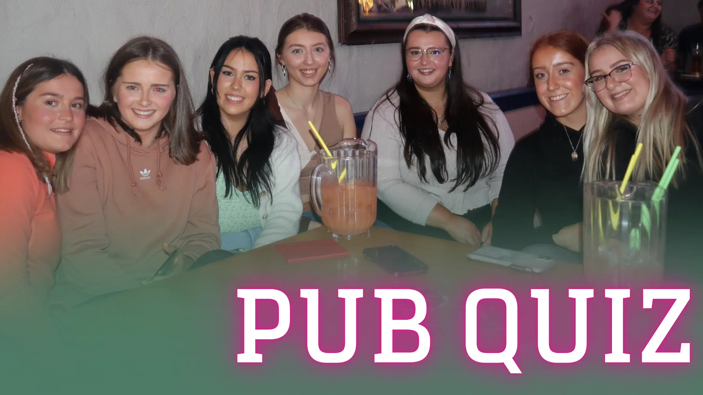 A group of smiling young women sitting at a table with drinks, enjoying a night out at a pub quiz event. The words 'PUB QUIZ' are boldly displayed at the bottom in large white and pink letters.